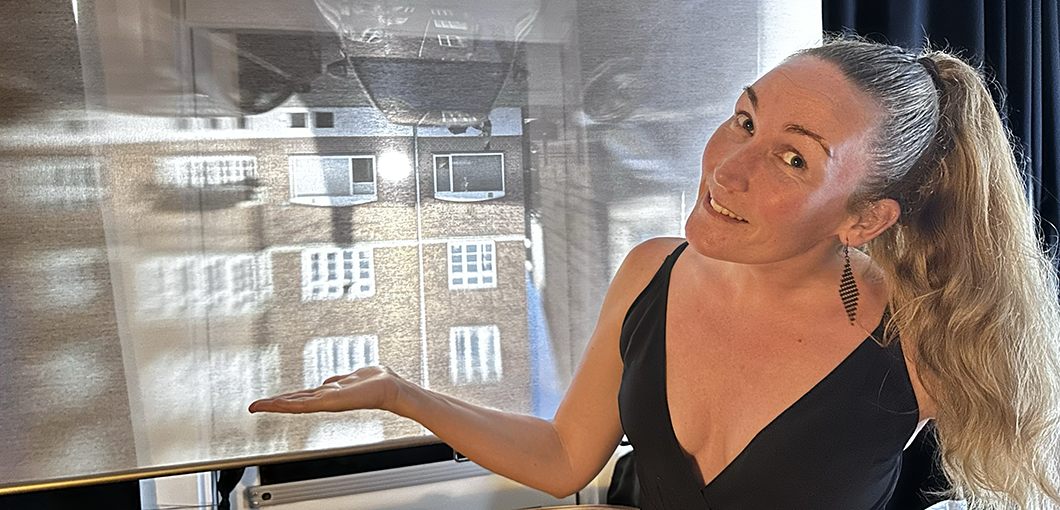 Christina Nahnsen, known as MissBlindSpot, is standing and pointing at a canvas with an upside-down section of a building and street, illustrating what a 'camera obscura' is and how it functions.