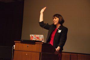 Lauren at a podium with her hand raised to wave to audience