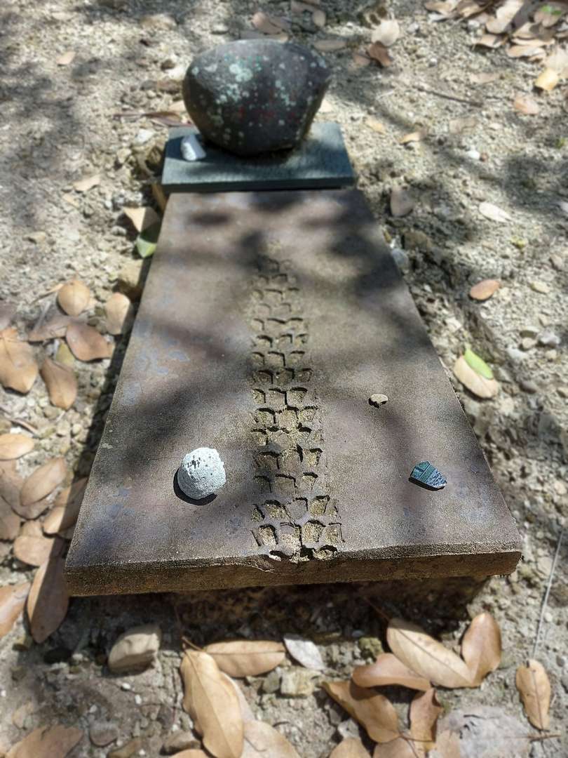 Eine Grabplatte aus Beton mit eingeprägter Reifenspur. Ein rundlicher, dunkler Stein ziert das Kopfende.