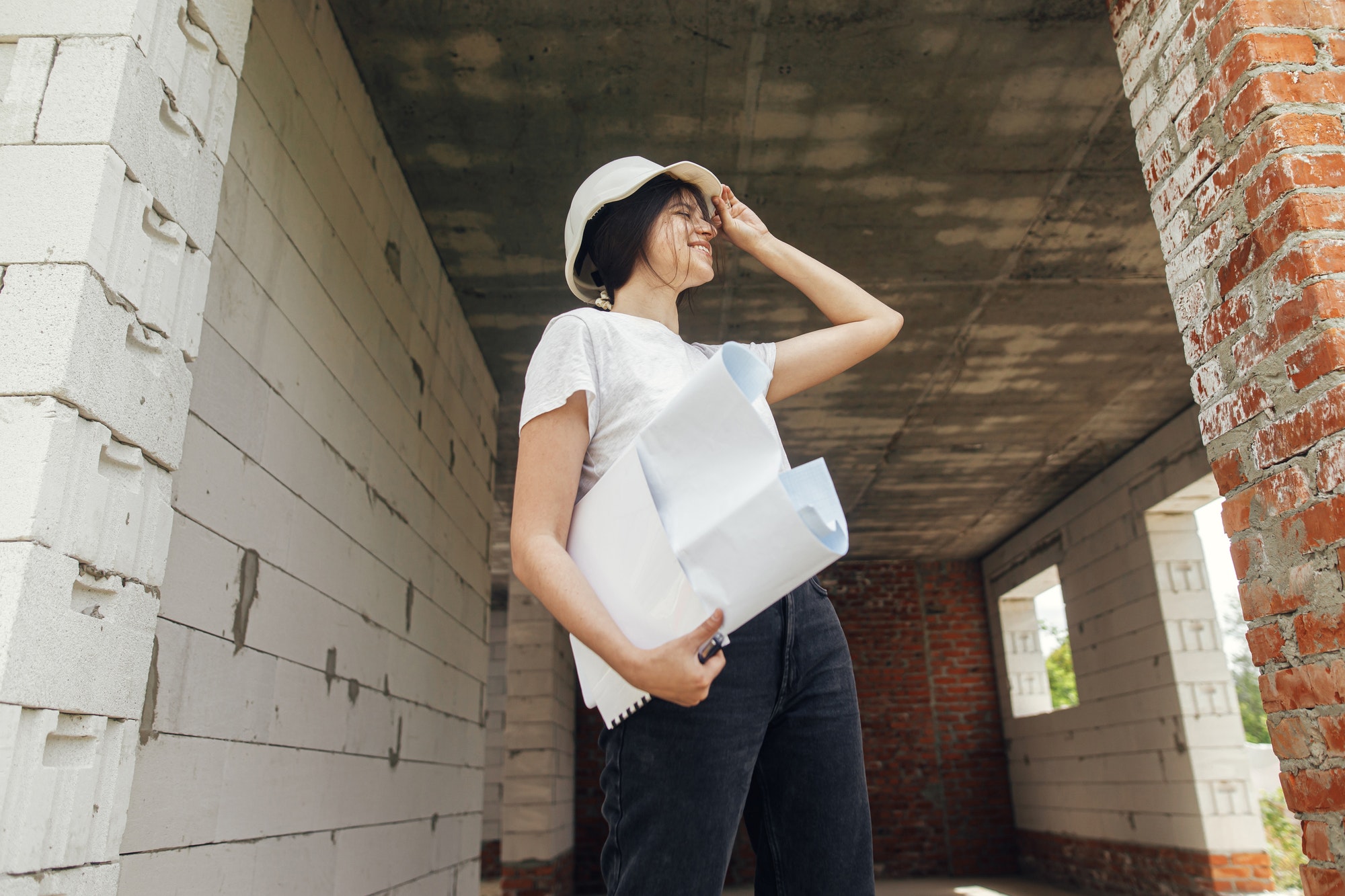 Stylish happy woman architect in hardhat checking blueprints in building house at construction site