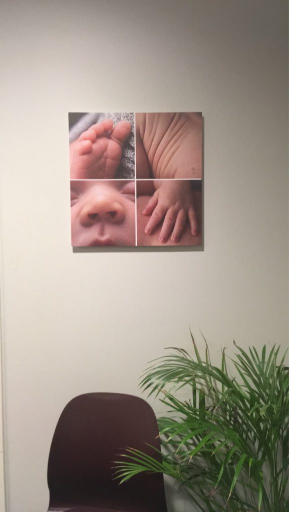 Et sammensatt bilde av babydetaljer henger på veggen over en stol og en plante på venterommet.