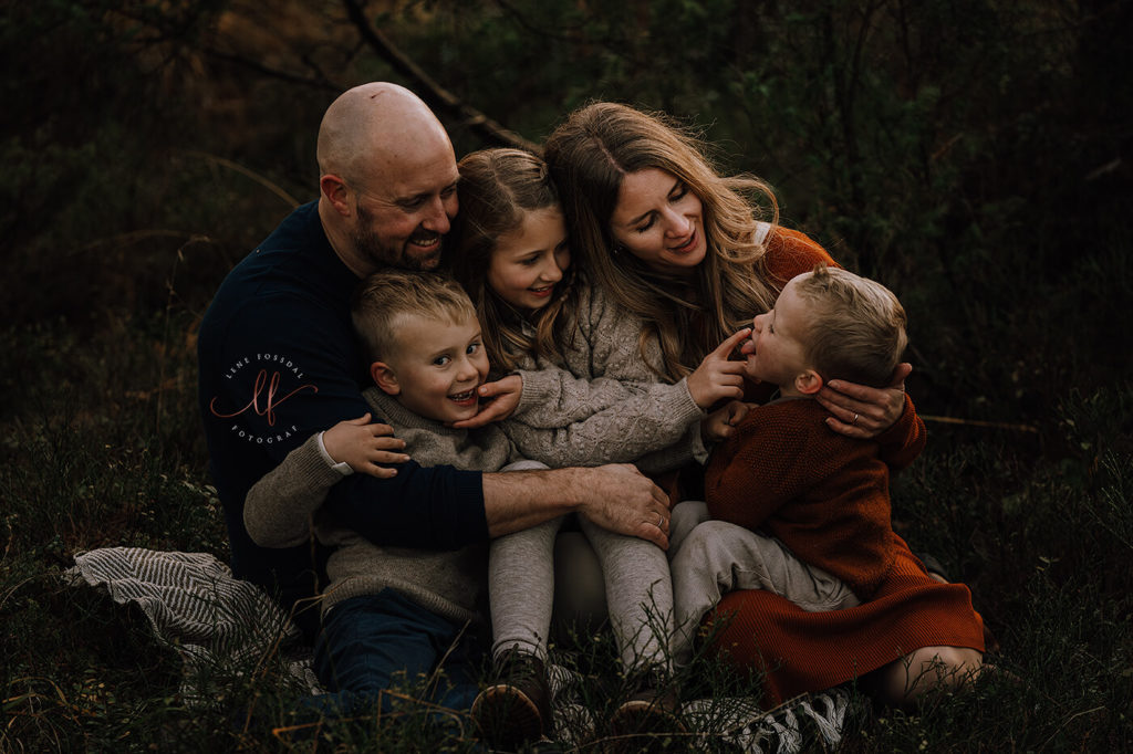 En familie sitter samlet i skogen. De er vendt mot hverandre, smiler og tøyser.