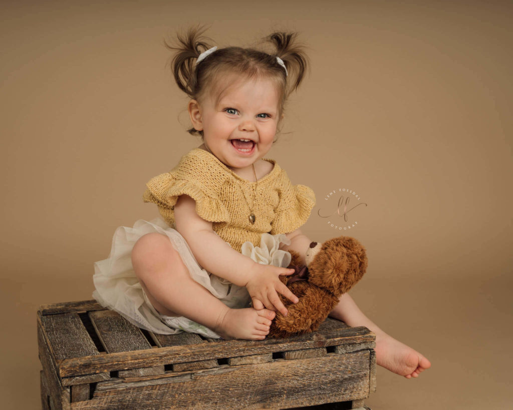 En jente på rundt et år sitter på en trekasse. Hun har musefletter og en gul kjole på, og smiler bredt mot kamera.