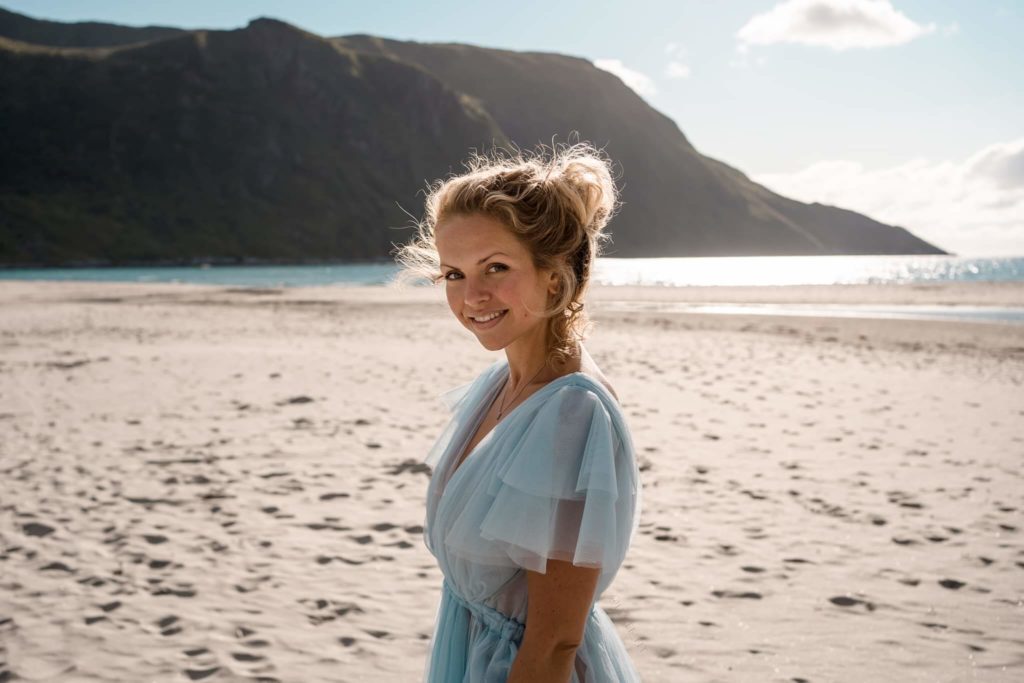 Lene Fossdal har på seg blå kjole på en strand. Bak henne er det et fjell som går ned i havet. Det er sol. 