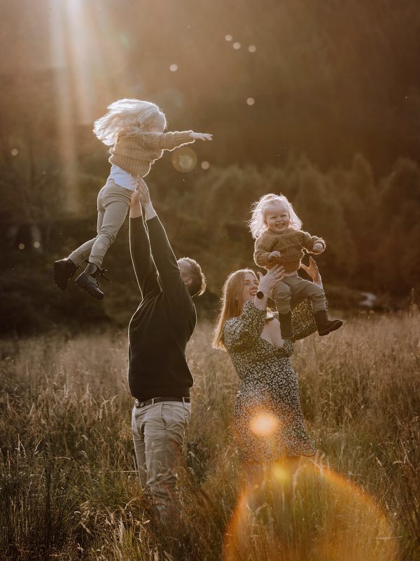 Et familiebilde tatt ute. Mor og far står med ryggen mot hverandre mens de kaster hver sin datter opp i luften. Det er i solnegang og bildet har varme toner.