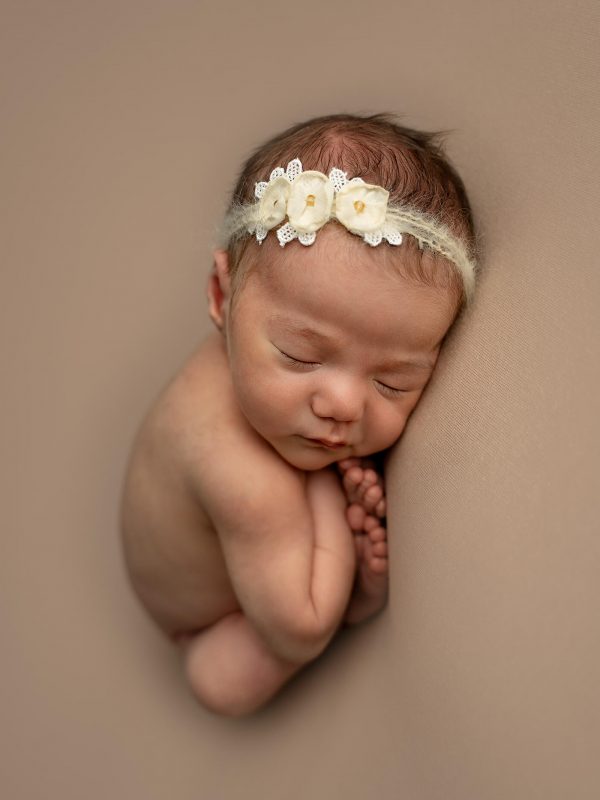 Dette er en jentebaby som ligger i såkalt "tacopose". Bildet er stående. Hun har hårbånd på hodet, mørkt hår og ligger sammenkrøllet med tærne nesten under haken.