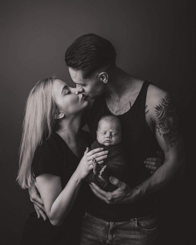En nyfødt baby holdes mellom far og mor som kysser. Bilde er tatt i studio, og er svarthvitt.