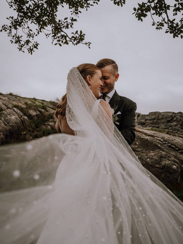 Brukeparet Kjersti og Christian. Kjersti står med ryggen til kameraet, Christian står mot henne og holder rundt henne. Sløret blåser mot kamera.