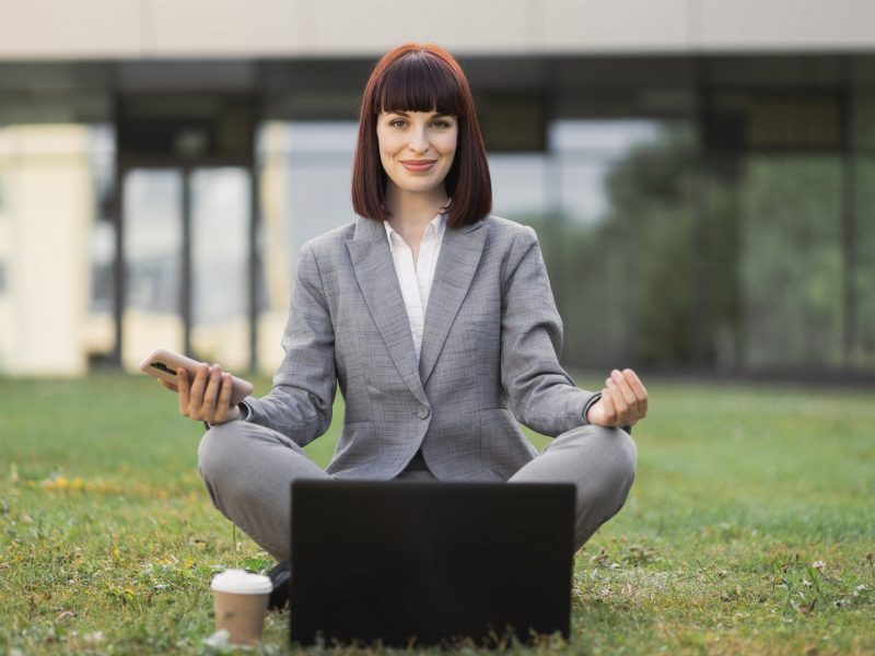 Mindfullness, no stress, free relief at work concept. Calm young woman relaxing meditating, sitting outdoors on green grass with laptop, practicing breathing yoga exercises and showing thumb up