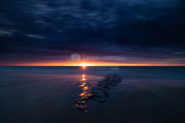 The sun sets and casts an orange glow over the sea and breakwater at the beach of Middelkerke, Flanders, Belgium