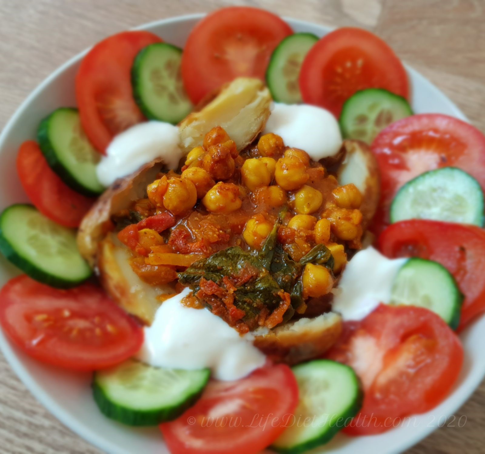Chickpea curry in a white bowl with baked potato, cucumber and tomato rings and yogurt.