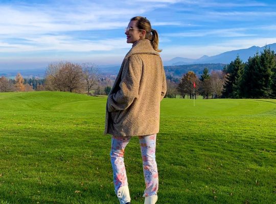 A coach for women, wearing a brown coat, floral pants, and white boots, stands on a grassy field at Lanserhof am Tegernsee, looking back. Trees, a red flag, and distant mountains are visible under a blue sky with clouds. coach