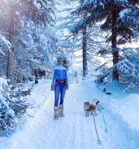 A coach for women stands in winter clothing on a snow-covered path in a forest near Seefeld in Tirol, Österreich, holding the leashes of two small dogs. Snow blankets the ground and trees beneath a clear sky. coach