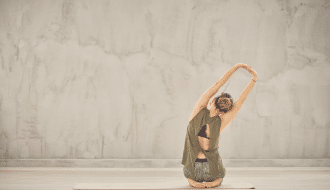 A person wearing a green tank top and grey shorts is kneeling on a yoga mat with arms extended and hands clasped overhead, embodying personal development. The background features a light grey textured wall.