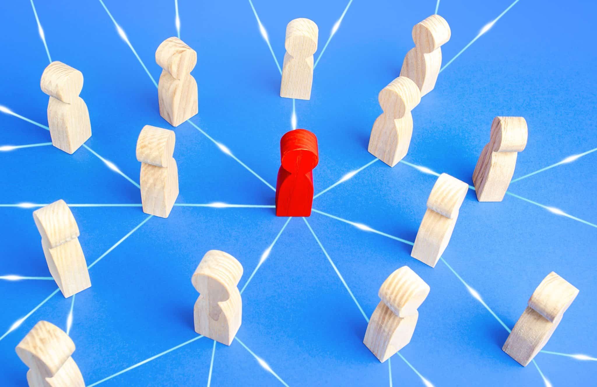 A group of eleven wooden peg figures arranged in a circular formation surrounds a single red wooden peg figure in the center. Lines connect the red figure to each of the surrounding figures, representing boundaries and connections, all placed on a blue background.