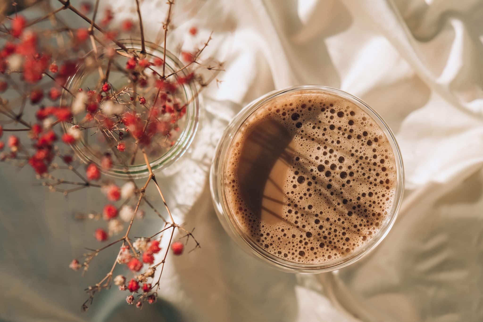 A glass of frothy coffee sits on a white fabric surface, casting shadows and showcasing light patterns. To the left, a vase holds delicate branches with small red and white flowers in focus, adding a soft contrast to the scene, gently illustrating the boundaries between simplicity and elegance.