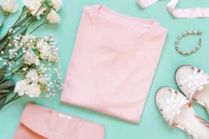 A flat lay image displays a neatly folded pastel pink t-shirt, a pair of white sandals, a pink clutch, a pearl bracelet, and a bouquet of white flowers arranged on a light turquoise surface—perfect for the stylish woman embracing personal development.