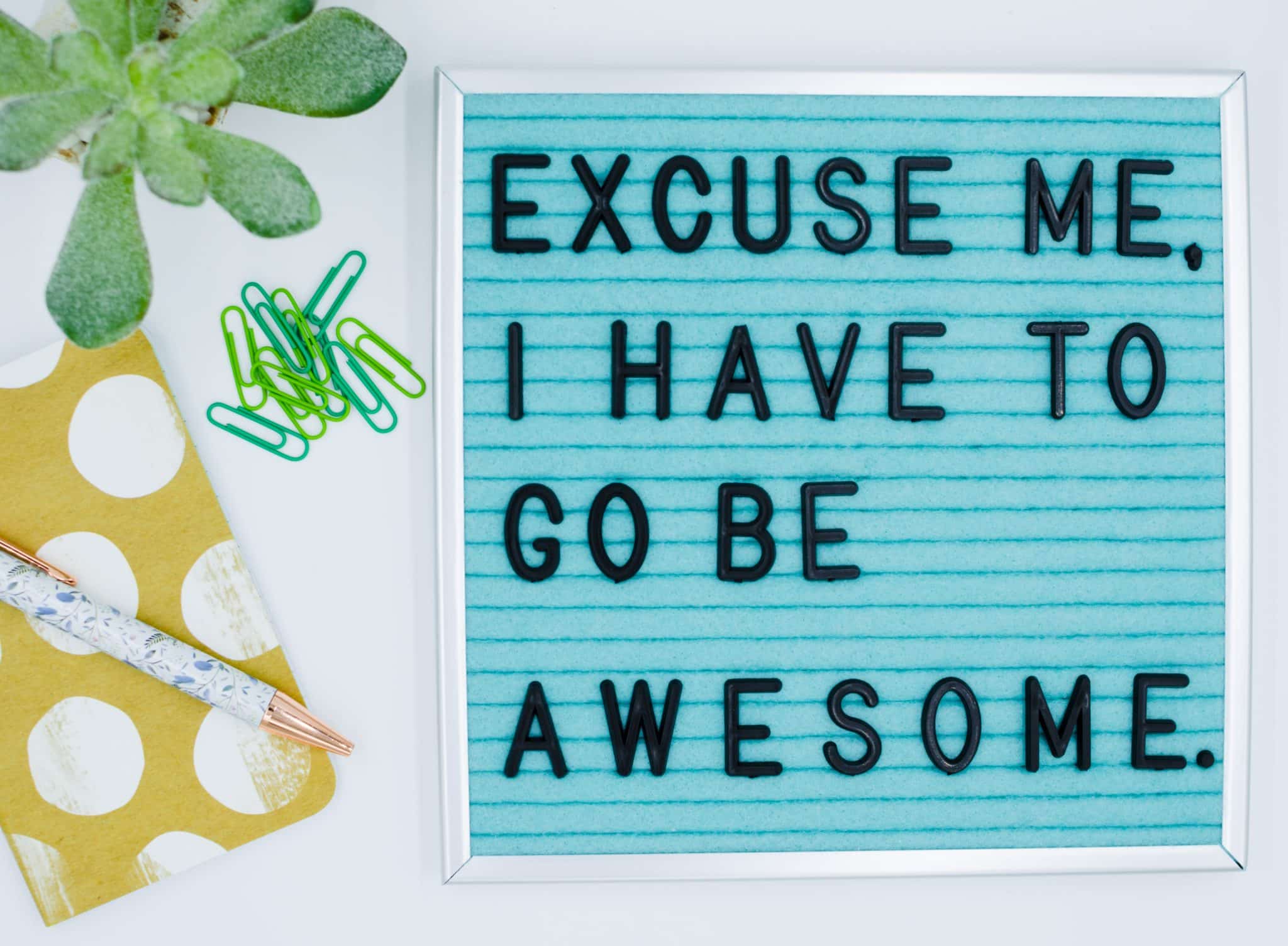 A letter board reads, "Excuse me. I have to go be awesome." The board has black letters on a light blue background and is surrounded by a succulent plant, green paper clips, a yellow polka-dot notebook, and two pens—the perfect setup for personal development and womens coaching inspiration.