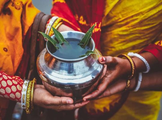 Three hands, adorned with colorful bangles and traditional clothing, hold a silver pot filled with water and green leaves. The focus is on the pot and the hands rather than faces, capturing a meaningful moment that could easily be guided by a coach for women, celebrating unity and cultural tradition. coach