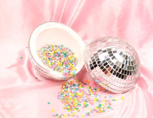 A broken disco ball lies on a pink satin surface. One half of the disco ball is filled with colorful confetti, which spills onto the fabric. The scene is vibrant and reflective, contrasting with the soft background.