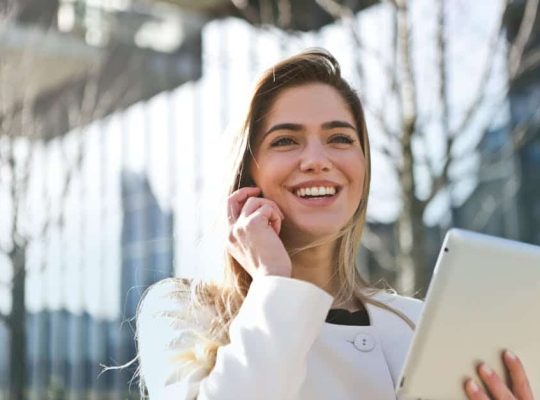 A coach for women, wearing a light-colored coat, stands outdoors smiling as she holds a tablet in one hand and touches her face with the other. In the background, glass buildings and bare trees add to the scene. coach
