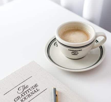 A cup of cappuccino on a saucer sits on a white table next to a closed notebook titled "The Gratitude Journal" and a silver pen—an inviting scene for any coach for women. White curtains are visible in the background, completing this calm and inspiring workspace. coach