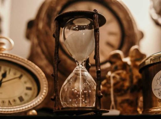 A close-up of an hourglass with sand running is surrounded by an antique clock on the left, a blurred figurine in the background, and a jar labeled "vintage" on the right—an evocative scene that could symbolize how a coach for women helps clients make the most of their time and treasures from the past. coach