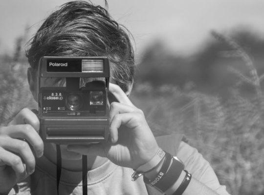 A coach for women holds a vintage Polaroid camera up to her face, taking a photo outdoors. The image is in black and white, with the background blurred. She wears several wristbands on her wrist. coach