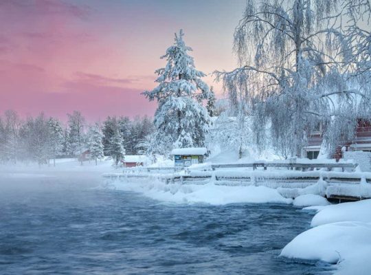 A snowy riverside scene at sunset, beautifully captured by a talented coach for women, features icy trees and a wooden fence along the riverbank. Snow-covered buildings nestle nearby while mist rises from the dark water under a softly glowing pink and blue sky. coach