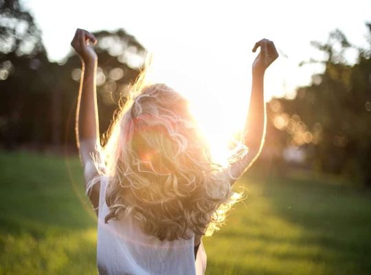 A coach for women with wavy blonde hair stands outside with arms raised, facing the sun. The sunlight creates a bright glow and lens flare, while green grass and trees are visible in the background. coach