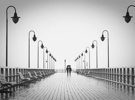 Two people walk under an umbrella on a wet, wooden pier lined with empty benches and evenly spaced street lamps, the overcast skies providing a calm backdrop—a tranquil scene that might set the perfect reflective mood for conversations with a coach for women. coach