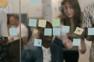 People are collaborating in an office setting, seen behind a glass wall covered with sticky notes in various colors. Some notes have markings on them. The individuals appear to be discussing or brainstorming, with focused expressions.