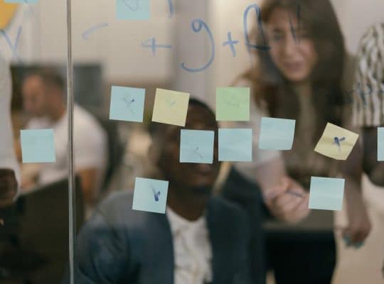 People are collaborating in an office setting, seen behind a glass wall covered with sticky notes in various colors. Some notes have markings on them. The individuals appear to be discussing or brainstorming, with focused expressions.