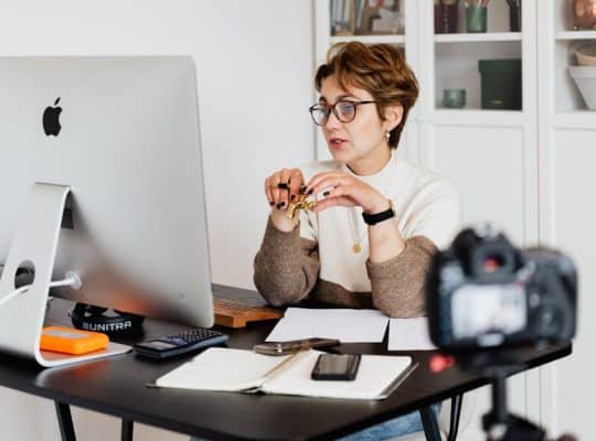 A coach for women, wearing glasses and a sweater, sits at a desk with a computer, notebook, and smartphone. A camera perched on a tripod is situated in the foreground, while shelves filled with various items are visible in the background. coach