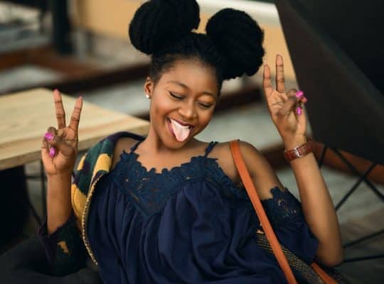 A person sits in a relaxed pose, sticking their tongue out playfully while embodying the spirit of a confident coach for women. Dressed in a dark blue top, they hold up peace signs with both hands, and their hair is styled in two large buns. coach