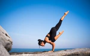A coach for women demonstrates an advanced yoga pose on a rocky surface, dressed in athletic clothing. She balances skillfully on her hands, with one leg extended upward and the other bent, set against the backdrop of a clear blue sky and the ocean. coach