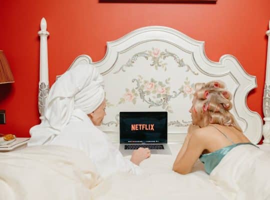 Two people are lying in bed, facing a laptop with the Netflix logo on the screen. One person, possibly a coach for women, has a towel wrapped around their head, while the other has curlers in their hair. The scene is set against a decorative headboard and a vibrant red wall. coach