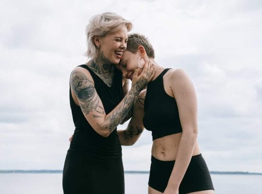 Two people—both wearing black sleeveless tops and shorts—stand outside near the water under a cloudy sky. One, who appears to be a coach for women, is smiling and embracing the other person, who is leaning in and smiling as well. Visible tattoos add a touch of personality to their warm interaction. coach