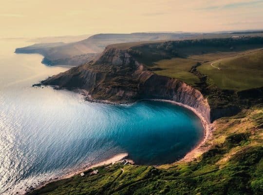 A coach for women immerses her clients in empowerment as they observe an aerial view of a coastal landscape, featuring a curved bay and cliffs. The deep blue sea is bordered by rugged cliffs and green hills, all beautifully lit by soft sunlight that creates a tranquil and scenic atmosphere. coach