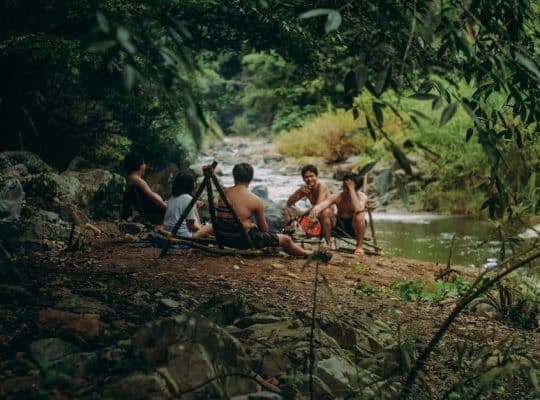A group of individuals, guided by a coach for women, sits on the ground near a riverbank, surrounded by trees. Some are shirtless. They appear to be engaged in conversation, with camping gear and a tripod nearby on the rocky terrain. coach