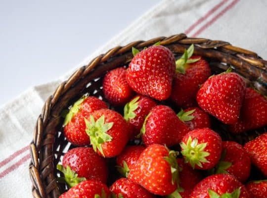 A wicker basket filled with ripe, red strawberries, perfect for a coach for women to enjoy during a break, is placed on a white cloth with red stripes. The strawberries, complete with their green leaves attached, create a vibrant scene set against a light gray background. coach