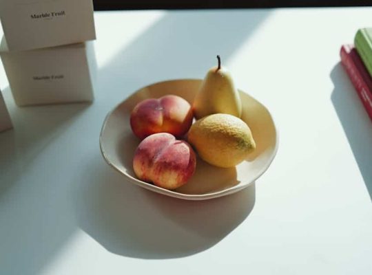 A beige plate holding two peaches, one pear, and one lemon sits on a white surface in the sunlight, with shadows cast across the table. Stacks of books or boxes are visible at the edges of the frame—a serene scene often found in the workspace of a coach for women. coach