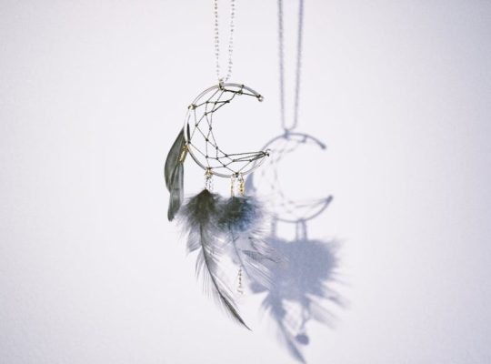 A crescent moon-shaped dreamcatcher with feathers and beads hangs against a white wall, casting its delicate shadow behind it—an inspiring sight for any coach for women seeking symbols of empowerment and dreams pursued. coach