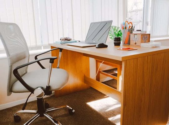 A modern office desk, ideal for a coach for women, features a mesh swivel chair, a closed laptop, mouse, notebook, pens, potted plant, mug, and various office supplies. The workspace is set near large vertical windows with blinds. coach