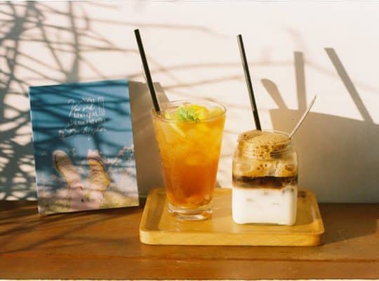 A glass of iced tea with lemon and mint and a jar of iced coffee with milk sit on a wooden tray next to a blue card or menu, bathed in sunlight and dappled shadows. This inviting scene creates the perfect backdrop for a coach for women to relax, reflect, and inspire her clients over refreshing drinks. coach