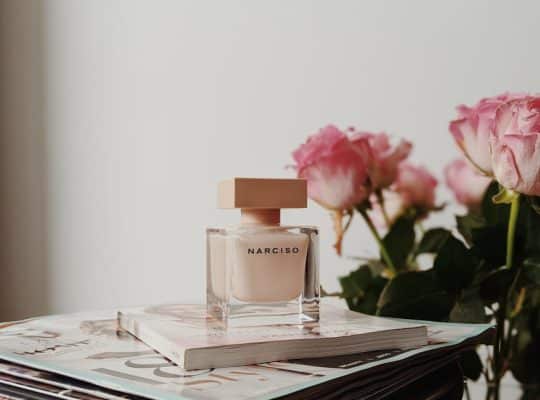 A bottle of Narciso perfume sits atop stacked magazines next to a bouquet of pink roses, styled as if curated by a coach for women, set against a plain white background. coach