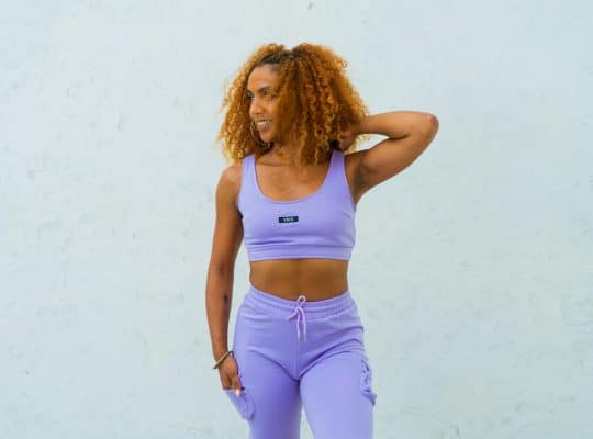 A coach for women with curly hair stands in front of a light-colored wall, wearing a matching lavender sports bra and pants. She has one hand behind her head and is looking to the side, smiling slightly. coach