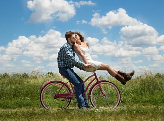 A man in a plaid shirt and jeans, who is a coach for women, rides a pink bicycle on a grassy path. He holds hands with a woman in a white dress and brown boots sitting on the handlebars. They are kissing under a blue sky with clouds. coach