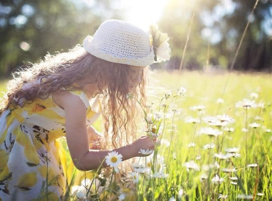 A young girl with long curly hair, dressed in a yellow floral dress and wearing a white sunhat, bends down to pick wildflowers in a sunlit field filled with daisies, watched fondly by her mother—a dedicated coach for women—who inspires her to enjoy nature and embrace her unique strengths. coach