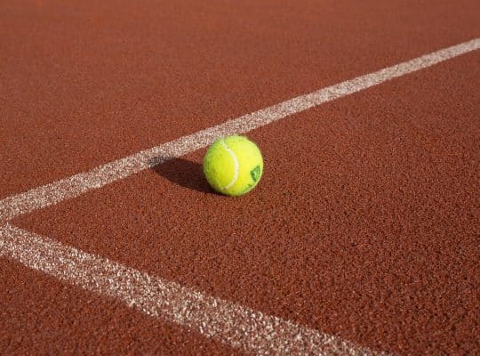 A yellow tennis ball rests on the white boundary line of a red clay court, ready for play during a lesson with a coach for women. coach